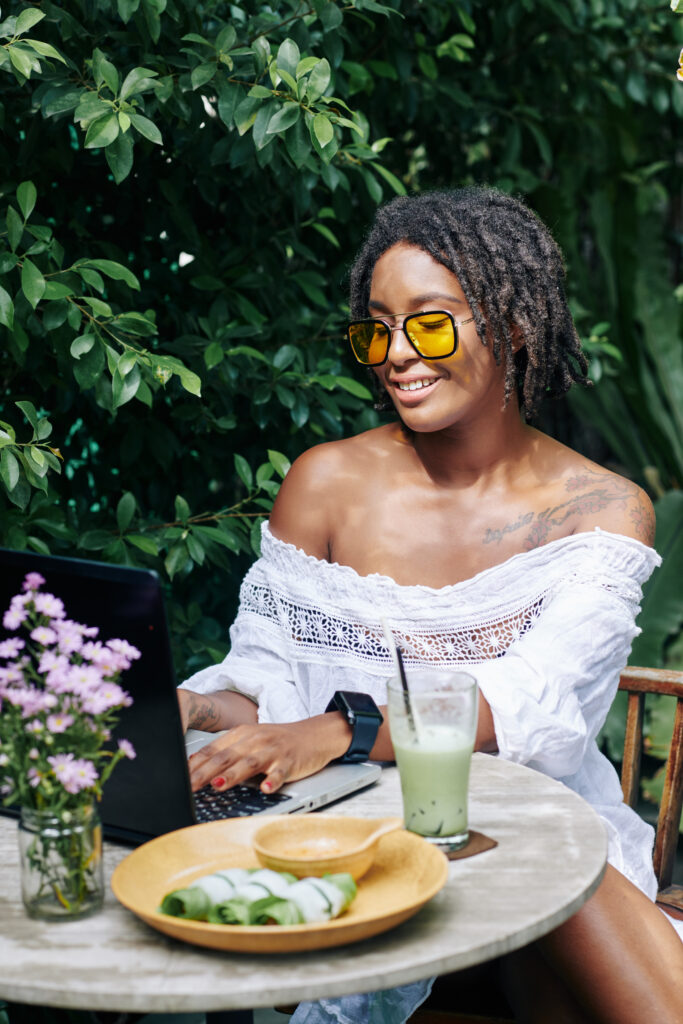 African smiling beautiful woman in sunglasses sitting at the table and typing on laptop computer during lunch in outdoor cafe